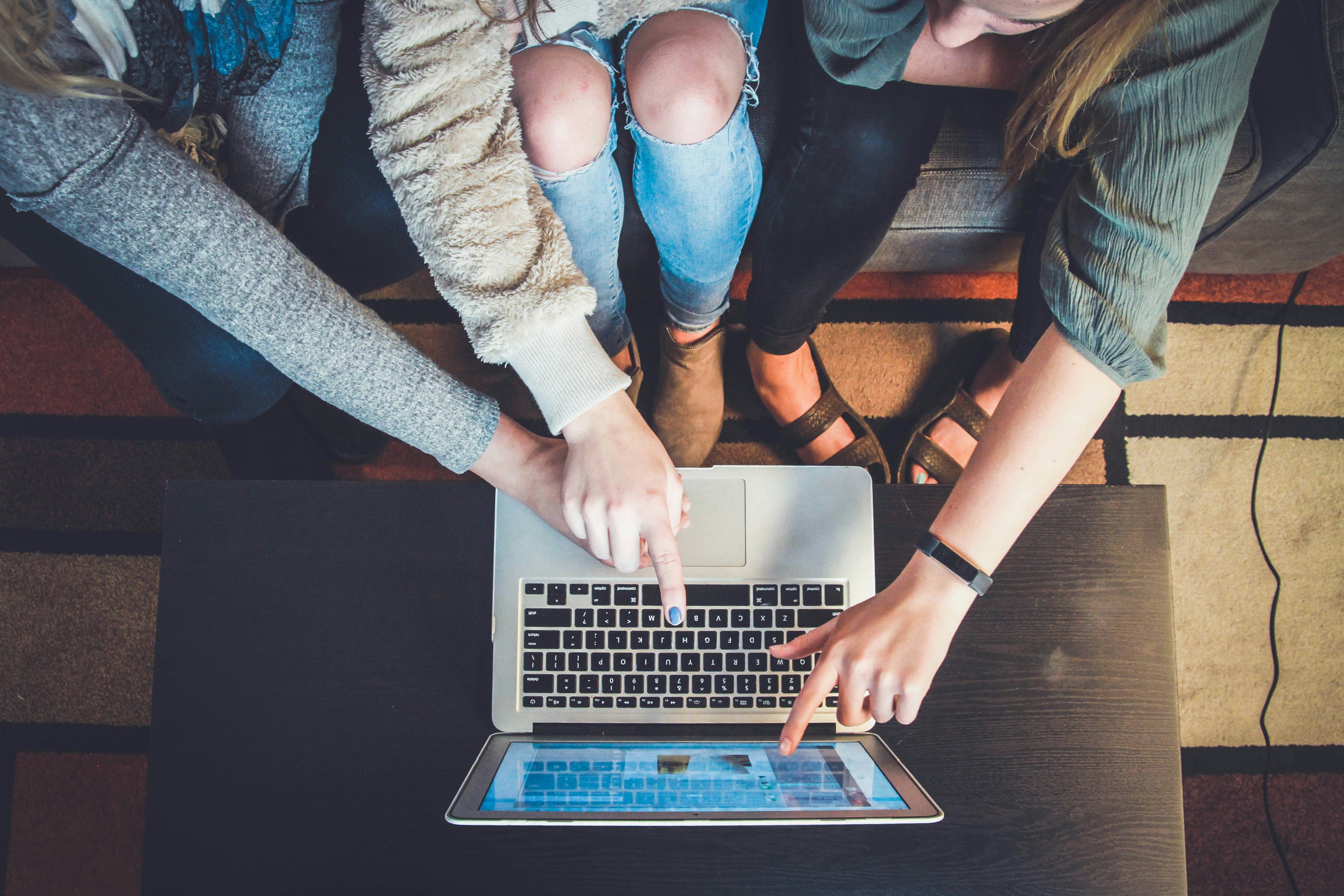 Drei Frauen zeigen auf einen Laptop der auf einem Couchtisch steht (Vogelperspektive)